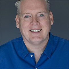 A man with blonde hair and blue shirt smiling.